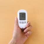A hand holding a blood glucose meter displaying 103 mg/dL on a neutral background.