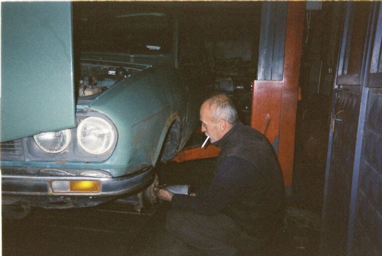 An elderly mechanic repairs a vintage car indoors, smoking a cigarette while working.