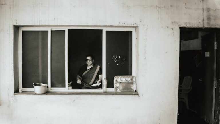 Woman in saree cooking indoors, viewed through window. Urban setting with visible utensils.