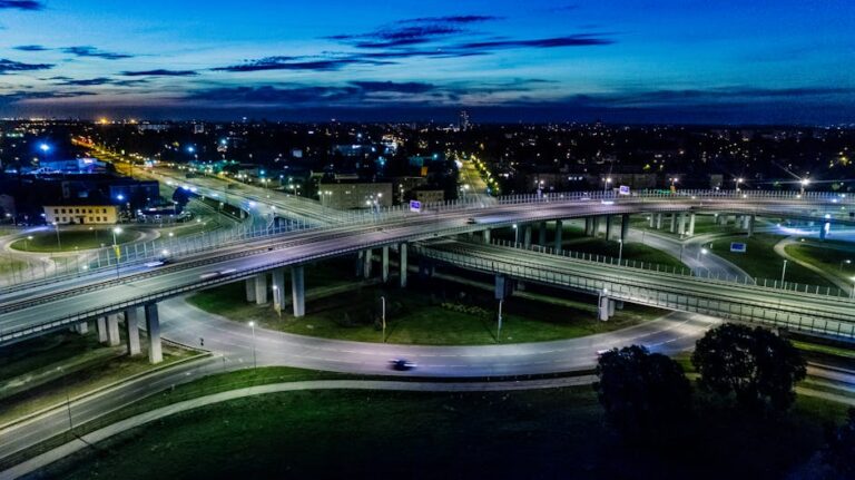 A dramatic cityscape featuring a modern flyover lit by vibrant city lights at night.