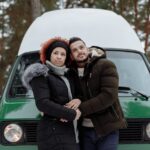 A couple stands embracing in front of a green van in a snowy forest during winter.