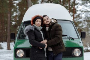 A couple stands embracing in front of a green van in a snowy forest during winter.