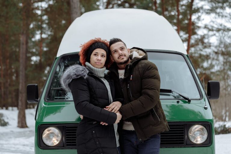 A couple stands embracing in front of a green van in a snowy forest during winter.