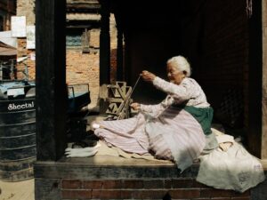 Senior woman weaving yarn with traditional tools on a sunny outdoor terrace.