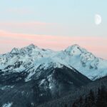 Majestic snow-covered mountain range with a rising moon and pink sunset sky.