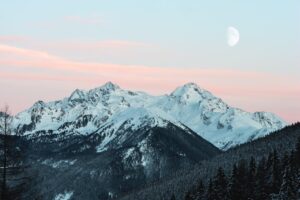 Majestic snow-covered mountain range with a rising moon and pink sunset sky.