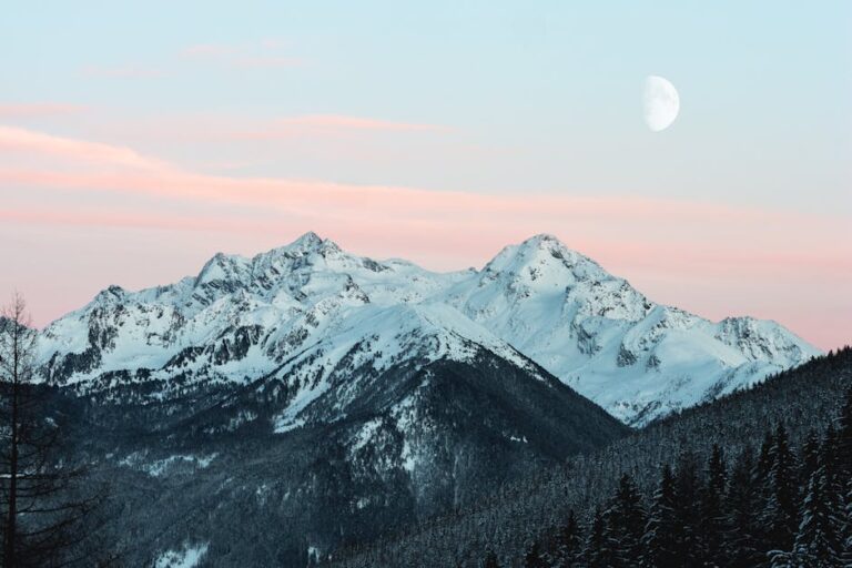 Majestic snow-covered mountain range with a rising moon and pink sunset sky.