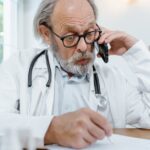Elderly doctor in office on phone call with patient, writing notes.