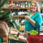 Senior woman shopping with a basket full of fresh produce in a grocery store, smiling at the camera.