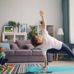A mother stretches on a yoga mat while her child plays with blocks nearby in their living room.