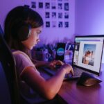 A young girl using a laptop with headphones in a dimly lit room, focusing on creative technology.