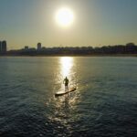 Person paddleboarding at sunset near city skyline, reflecting on calm sea.