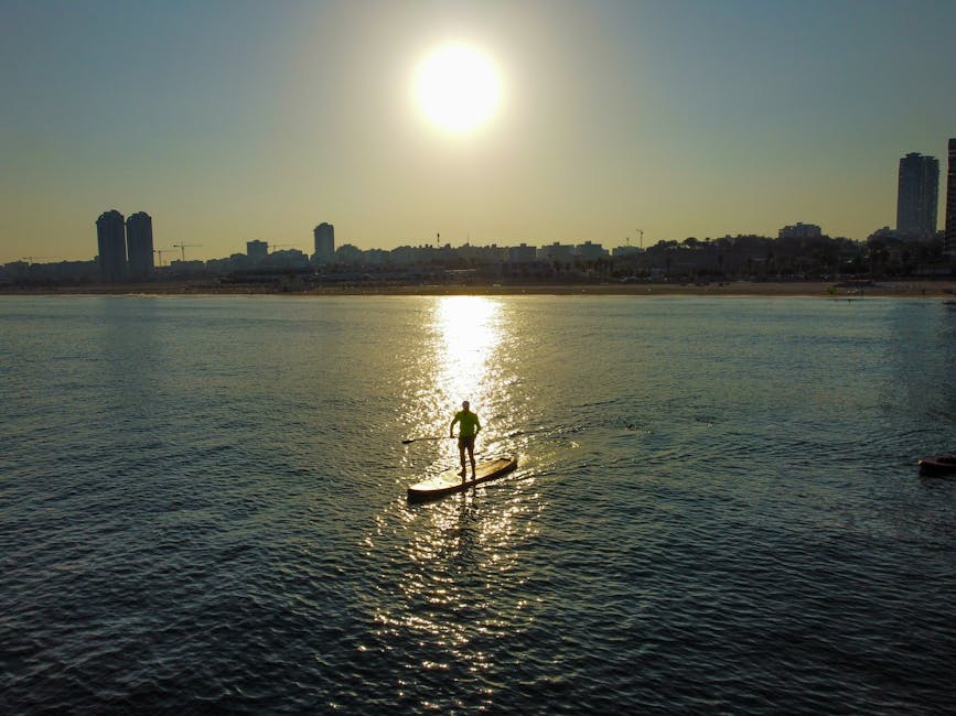 Person paddleboarding at sunset near city skyline, reflecting on calm sea.