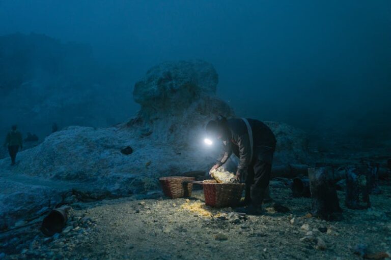 A solitary miner with headlamp gathers sulfur in baskets at a quarry during night.