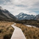 Wooden walkway leads through rugged landscape towards majestic snow-capped mountains.