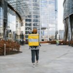 A delivery person with a yellow backpack walking through a modern urban cityscape, ready for service.