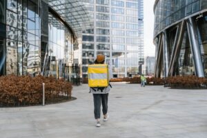 A delivery person with a yellow backpack walking through a modern urban cityscape, ready for service.