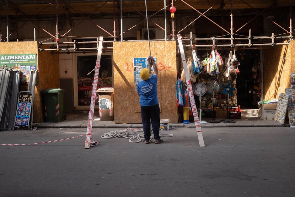 A construction worker standing on a street setting up scaffolding outside an urban storefront.