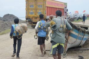 Three fishermen carrying fishing gear walking towards the harbor near fishing boats.